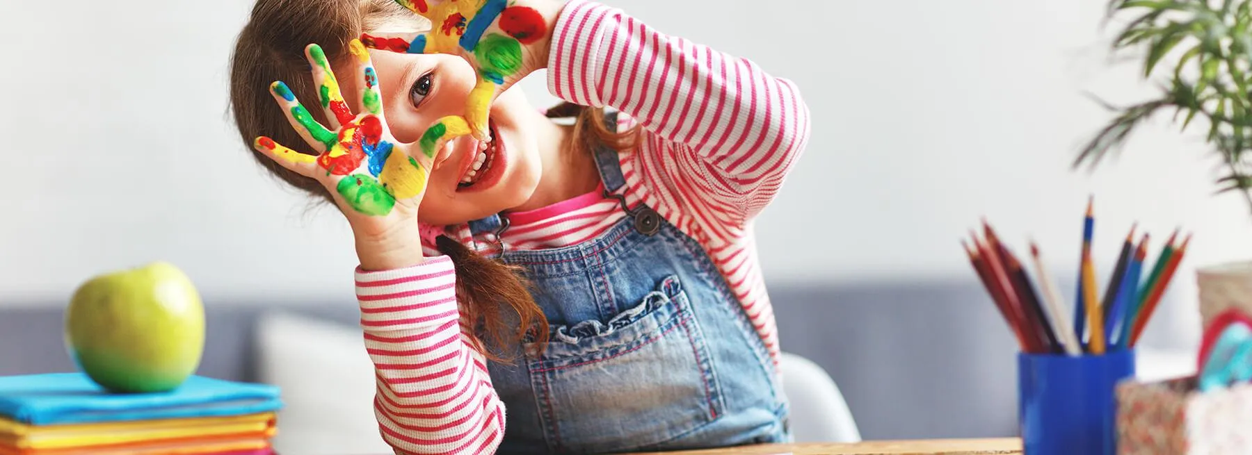 A young girl grinning through paint-covered hands at a craft table